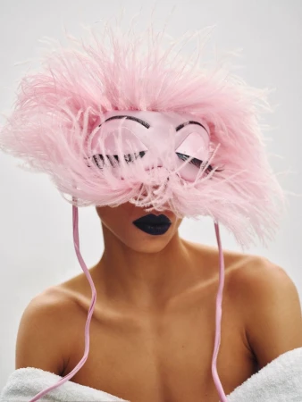 Model wearing a feathered pink mask with painted eyes and dark lipstick, photographed against a white background.