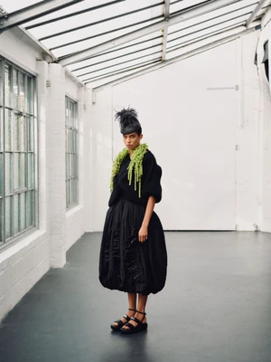 Model wearing a voluminous black dress and textured green scarf stands in a bright industrial studio space.