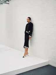 Model leaning against a white brick wall wearing a black jacket and matching skirt with white floral accents.