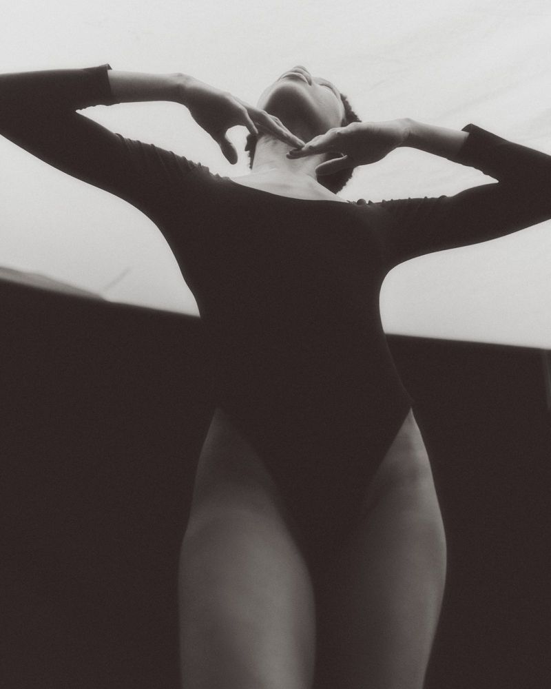 Model in a black leotard posing with arms raised, shot from a low angle in black and white.
