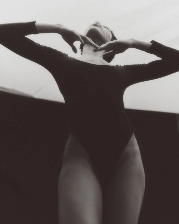 Model in a black leotard posing with arms raised, shot from a low angle in black and white.