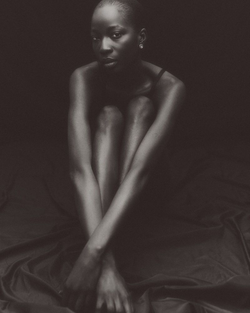 Monochrome portrait of a seated model with pulled-in knees against a dark draped backdrop.