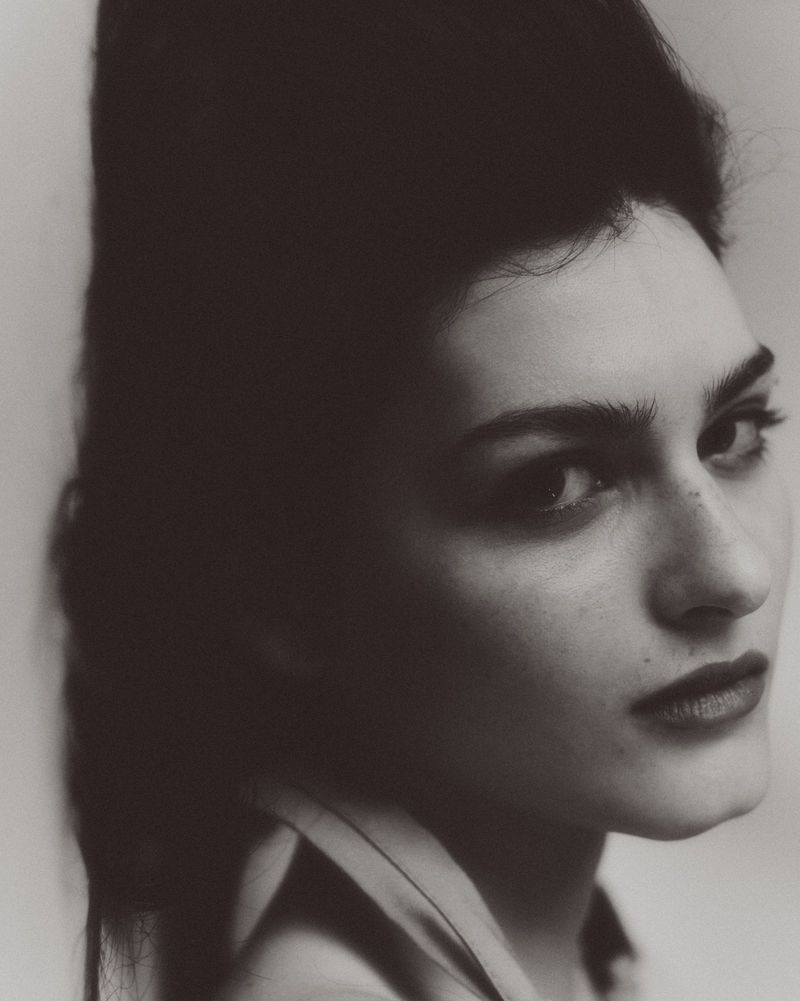 Close-up monochrome portrait of a model with voluminous dark hair and dark lipstick against a plain studio background.