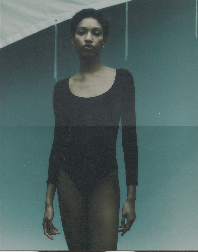 Studio shot of a slender adult woman with short hair posing in a black bodysuit against a cyan backdrop with hanging strings.