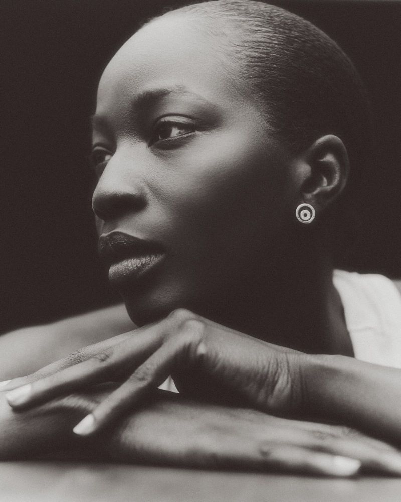Close-up monochrome portrait of a black woman resting her chin on her crossed hands, wearing a patterned stud earring.
