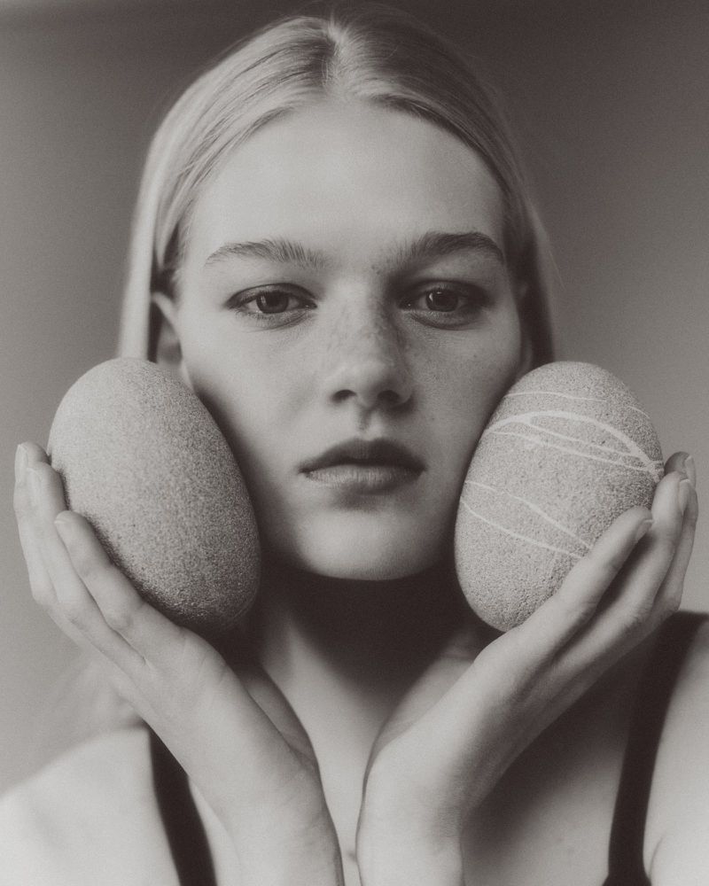 Portrait of a young woman holding smooth oval stones against her cheeks in a minimalist studio setting.