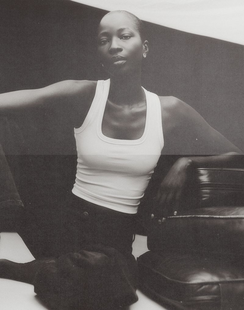 Monochrome portrait of a model wearing a white tank top seated beside dark leather luggage.