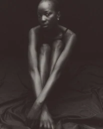 Monochrome portrait of a seated model with pulled-in knees against a dark draped backdrop.