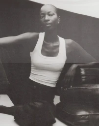 Monochrome portrait of a model wearing a white tank top seated beside dark leather luggage.