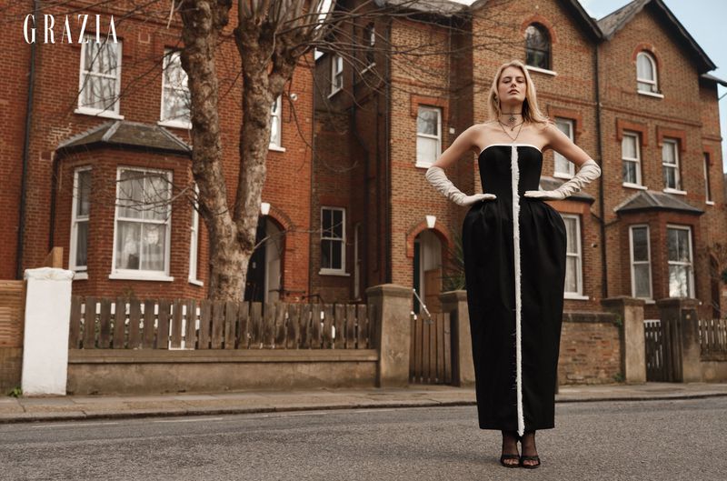 Model wearing a black and white strapless dress with long white gloves standing on a street in front of red brick residential buildings