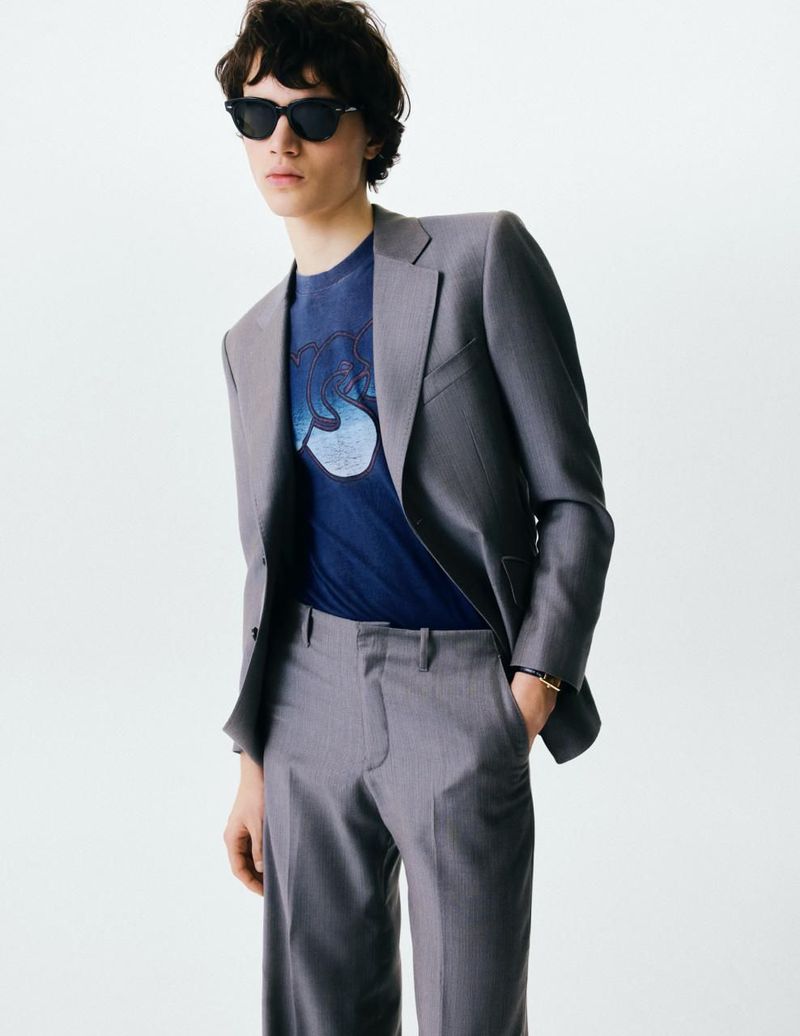 Model wearing a grey suit jacket and trousers over a blue t-shirt with sunglasses against a white background.
