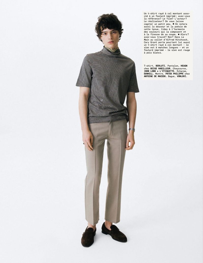A young male model standing against a white seamless background wearing a striped turtleneck t-shirt and grey trousers.