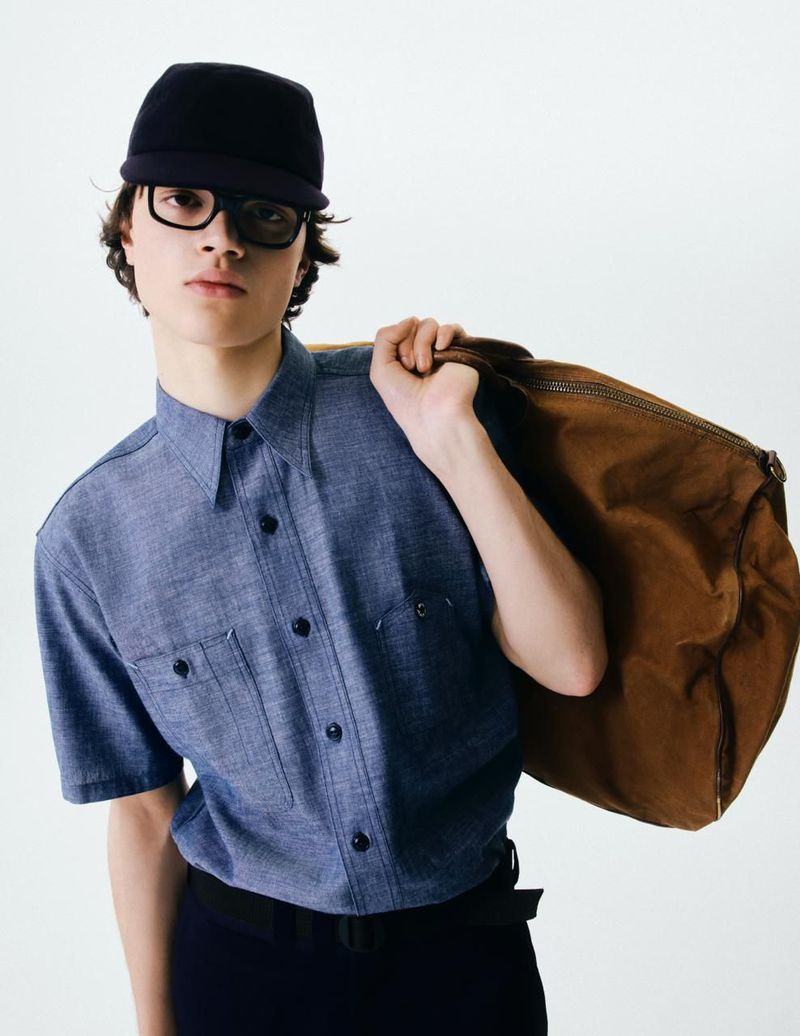 Young adult male poses in a photo studio wearing a dark blue button-down shirt, cap, glasses, and carrying a large brown duffel bag.