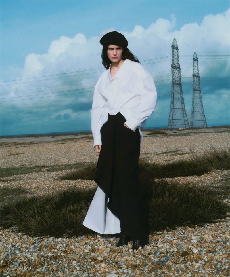Model wearing a black beret, white shirt, and black trousers standing outdoors in a flat, sparse landscape with industrial towers.