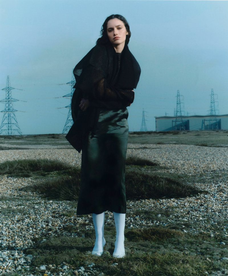 Model wearing dark clothing and white knee-high socks posing in a desolate landscape with industrial power pylons under a pale sky.