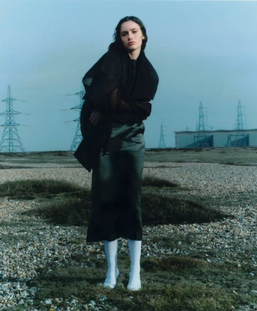 Model wearing dark clothing and white knee-high socks posing in a desolate landscape with industrial power pylons under a pale sky.