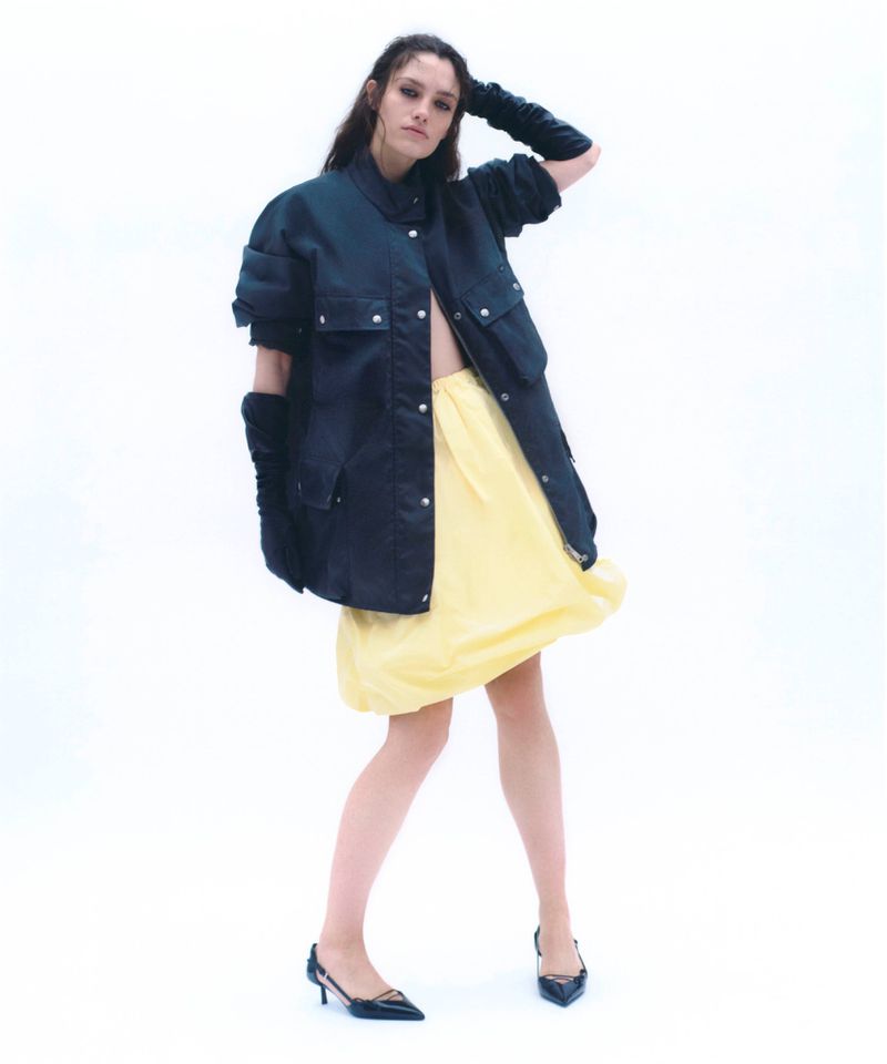 Model in black utility jacket and yellow skirt against a white studio background
