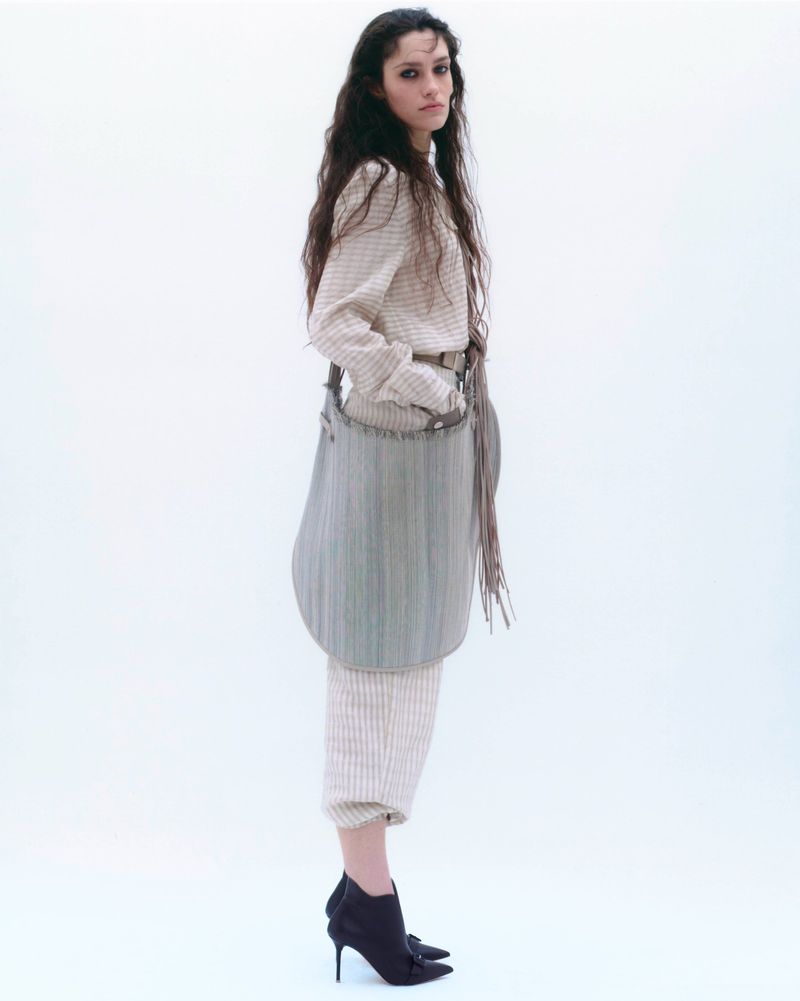 Model with long dark hair wearing a patterned beige outfit and holding a large grey handbag against a white background