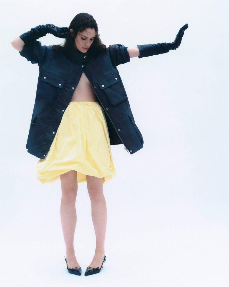 Model in a dark coat and yellow skirt wearing black gloves posing against a white background