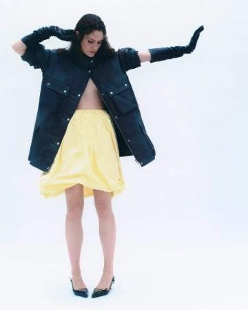 Model in a dark coat and yellow skirt wearing black gloves posing against a white background