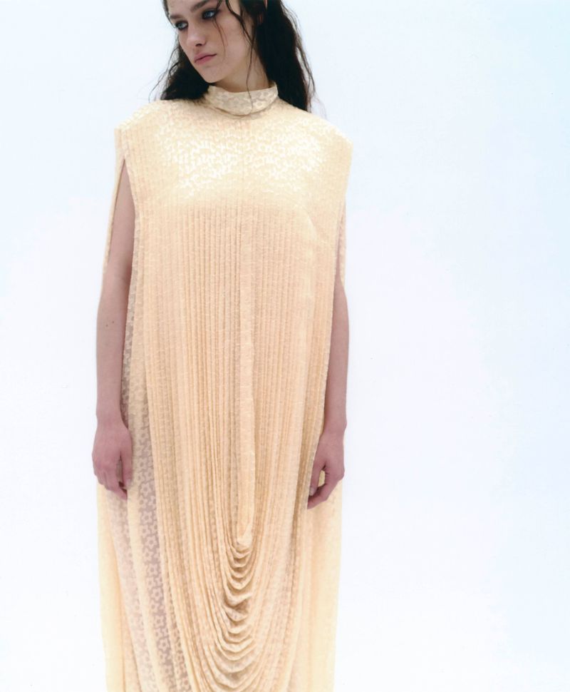 Model in a high-necked pale yellow draped dress standing against a seamless white background