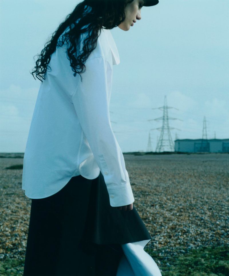 Model with dark curly hair wearing a white shirt and black skirt against a sparse coastal landscape with industrial power pylons