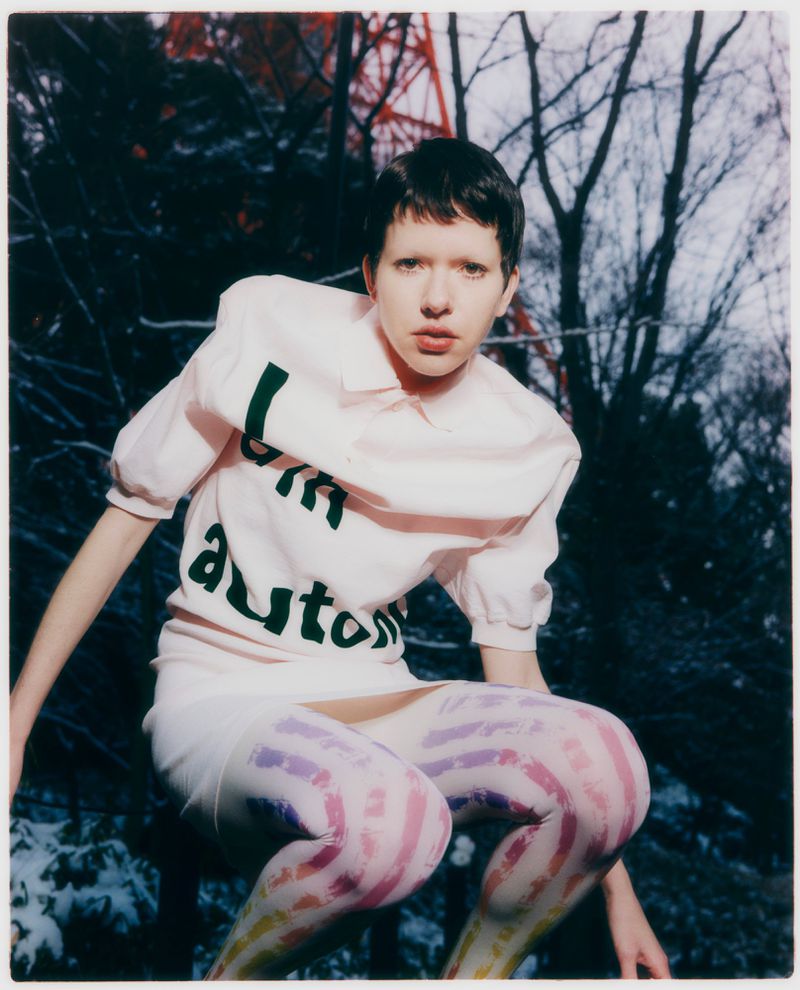 Model wearing a pink printed top and patterned tights outdoors against bare trees and a red tower structure.