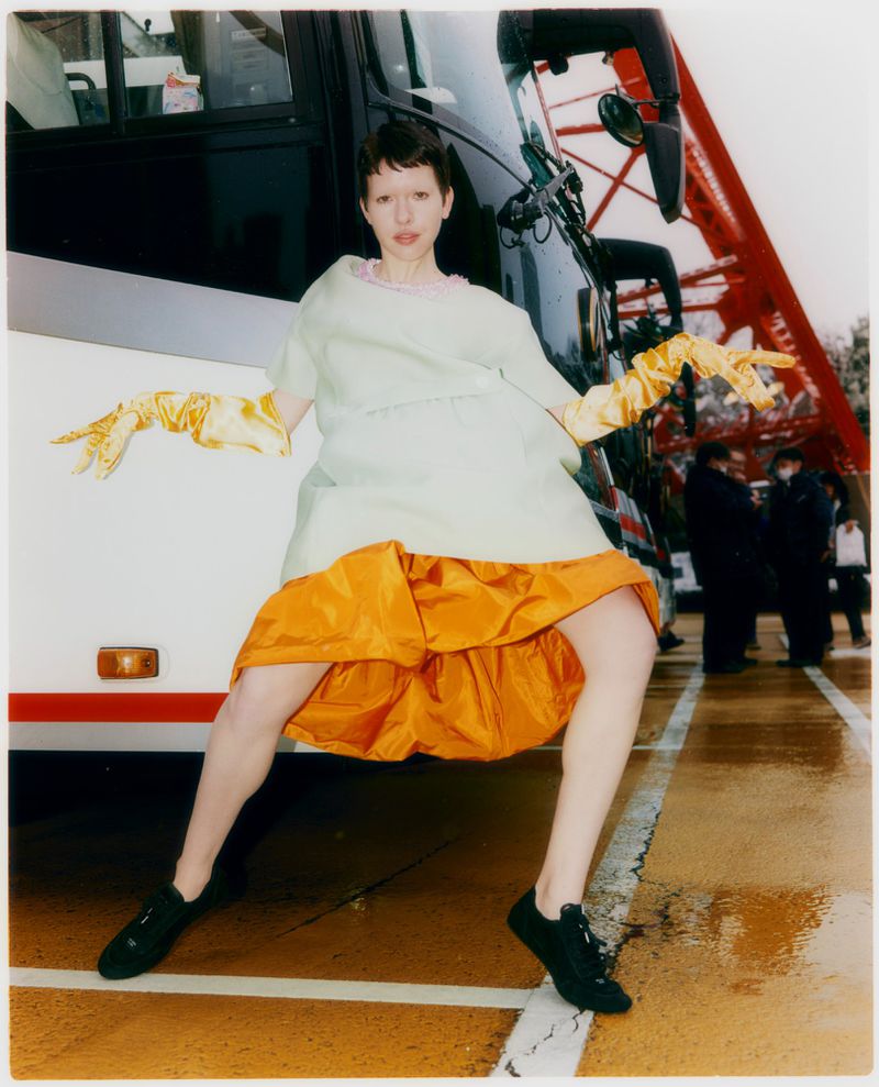 Model in a pale green top, orange ruffled skirt, and yellow gloves posing in front of a tour bus
