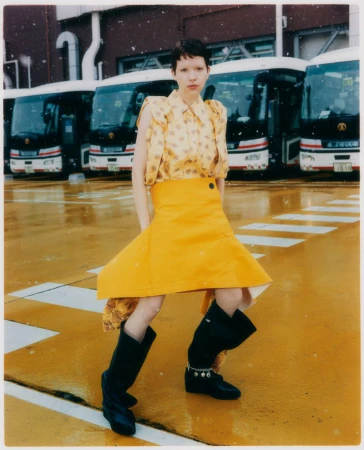 Model in yellow patterned blouse and bright yellow skirt with black boots standing before parked buses.