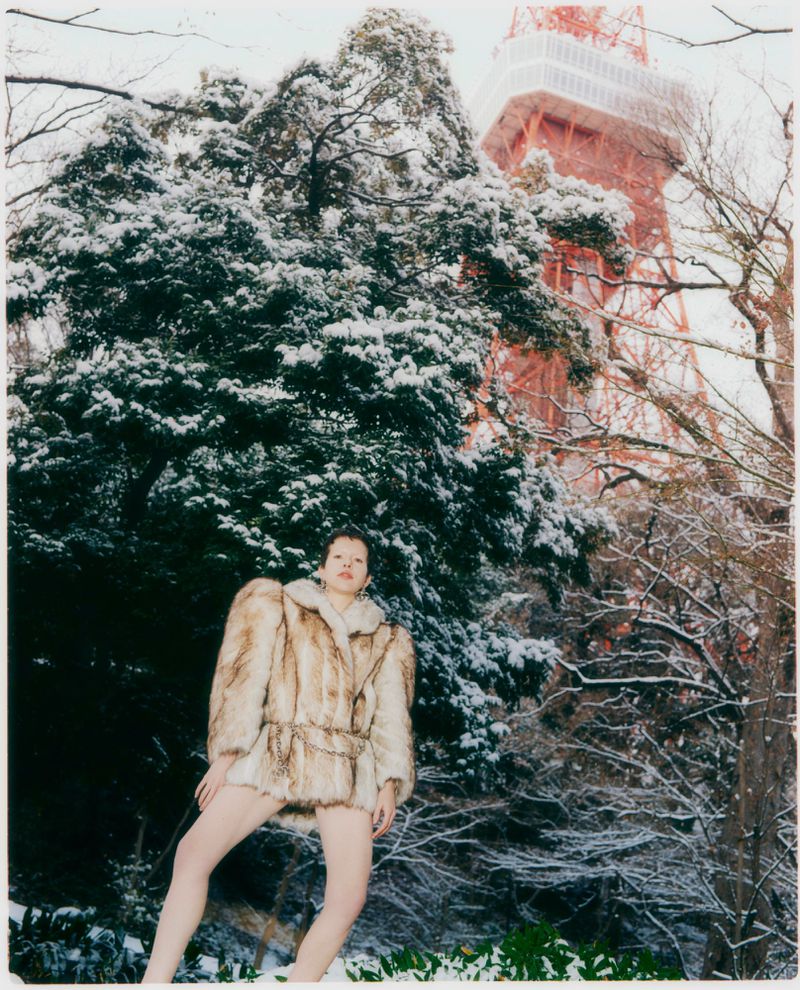 Model wearing a faux fur coat standing outdoors in a snowy park with a tall red tower in the background.