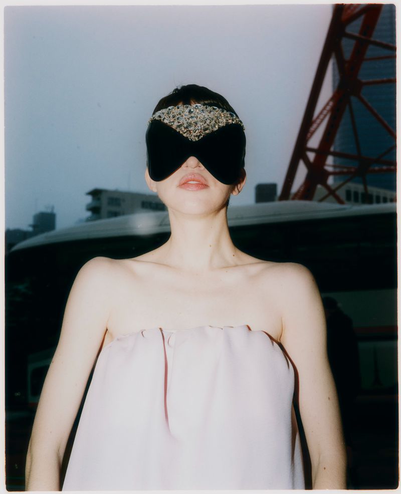 Model wearing a decorative black eye mask and strapless pale pink top outdoors with a red steel tower in the background.