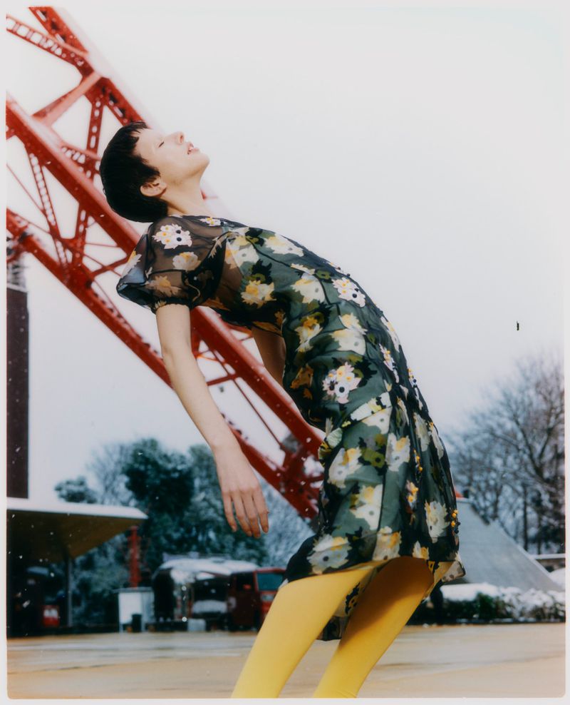 Model in a floral print dress and yellow tights leaning backward outdoors beneath a red architectural structure