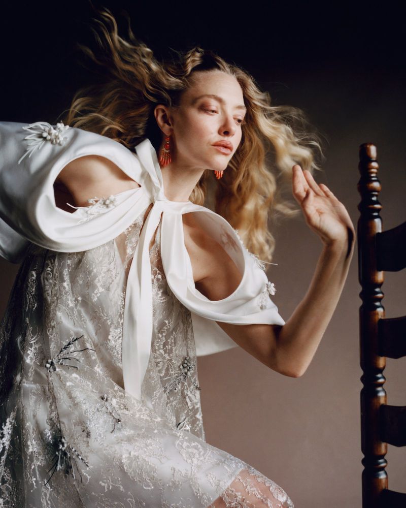 A blonde female adult in a white gown sits on a wooden chair in a studio with natural light. She wears coral earrings and poses.