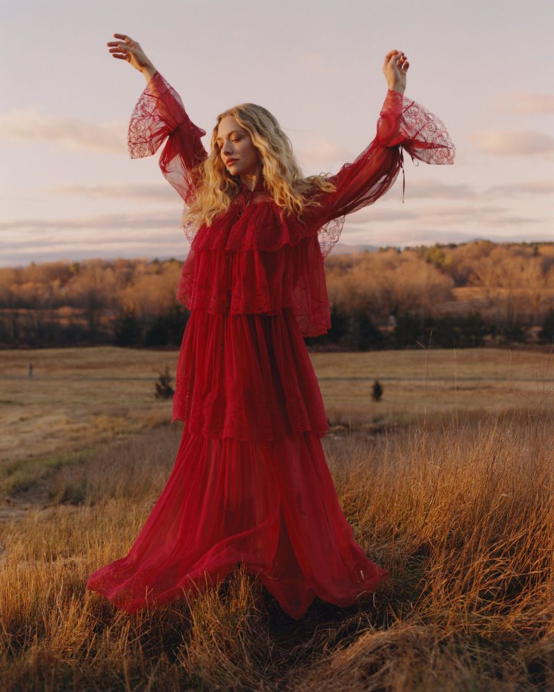 Amanda Seyfried wearing a long red lace dress with arms raised in an open grassy field during sunset.