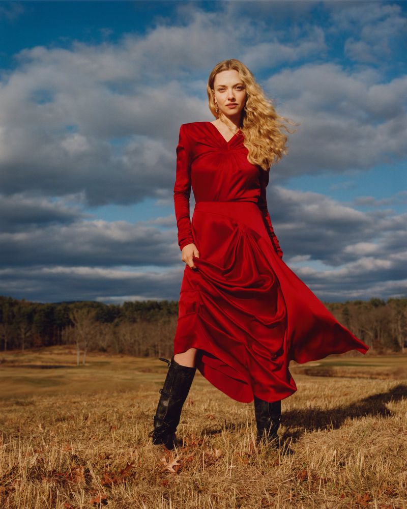 Amanda Seyfried wearing a red dress and tall black boots standing in an open field under a cloudy blue sky.