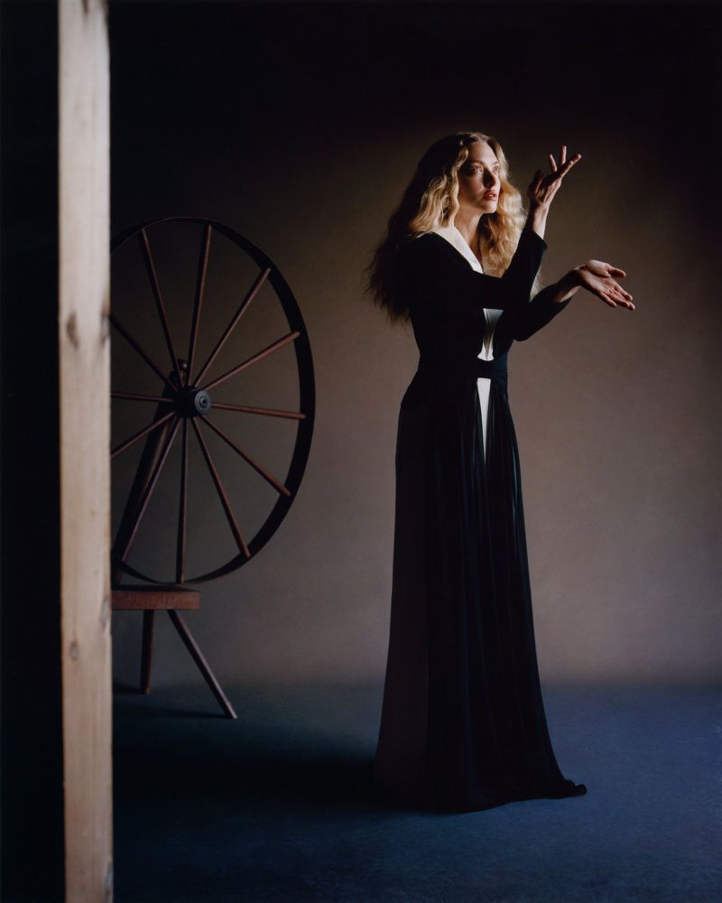 Amanda Seyfried wearing a long black and white dress posing beside a spinning wheel in an interior studio setting.