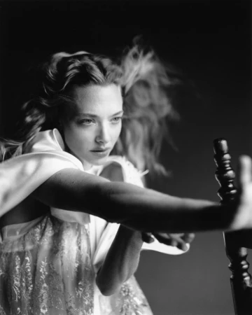 Portrait of Amanda Seyfried wearing a white dress extending her arm outward against a dark background
