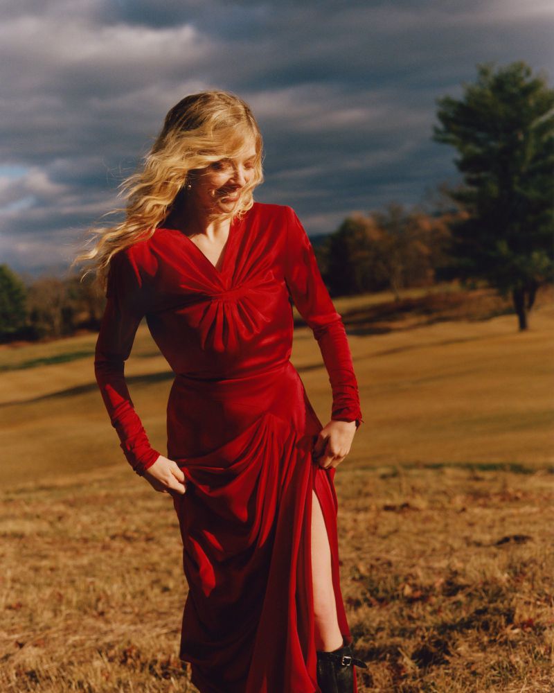 Amanda Seyfried wearing a long red dress walking outdoors in a grassy field under a cloudy sky for an American Vogue editorial.