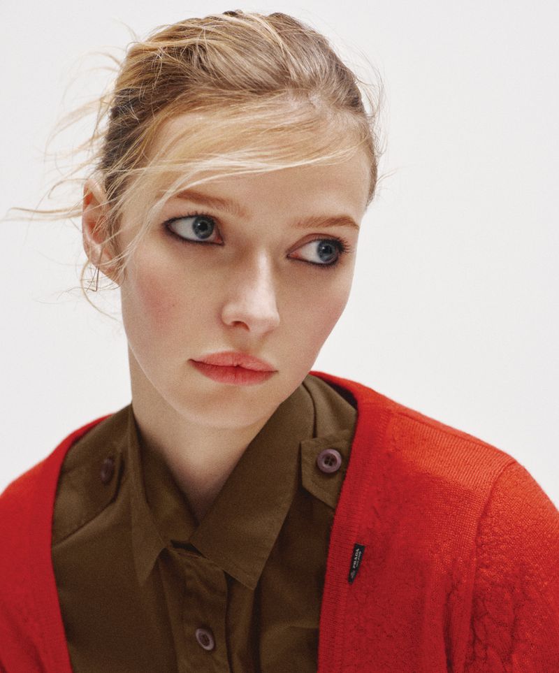 Close-up portrait of a young blonde model wearing an olive green button-down shirt and a red cardigan against a neutral white background.