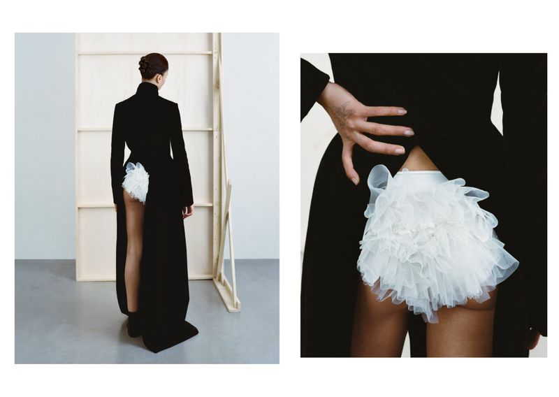 Woman in black dress with white ruffled detail poses in a photo studio. Fashion editorial with minimalist setting and neutral color palette.