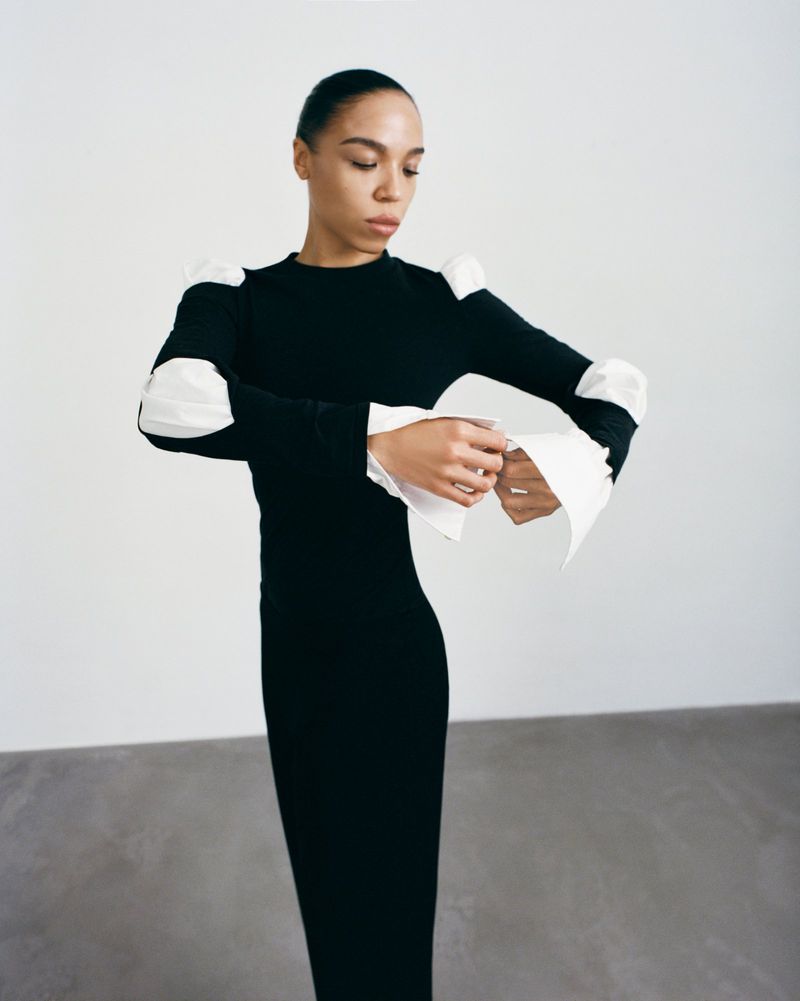 Elegant woman in long black dress with white sleeve details poses in a minimalist white studio, showcasing fashion and style.