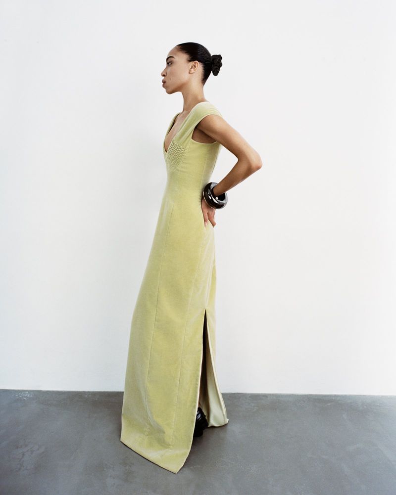 Elegant woman in a light green velvet gown, posing in a studio setting. She wears a large black bangle and has her hair in a bun.