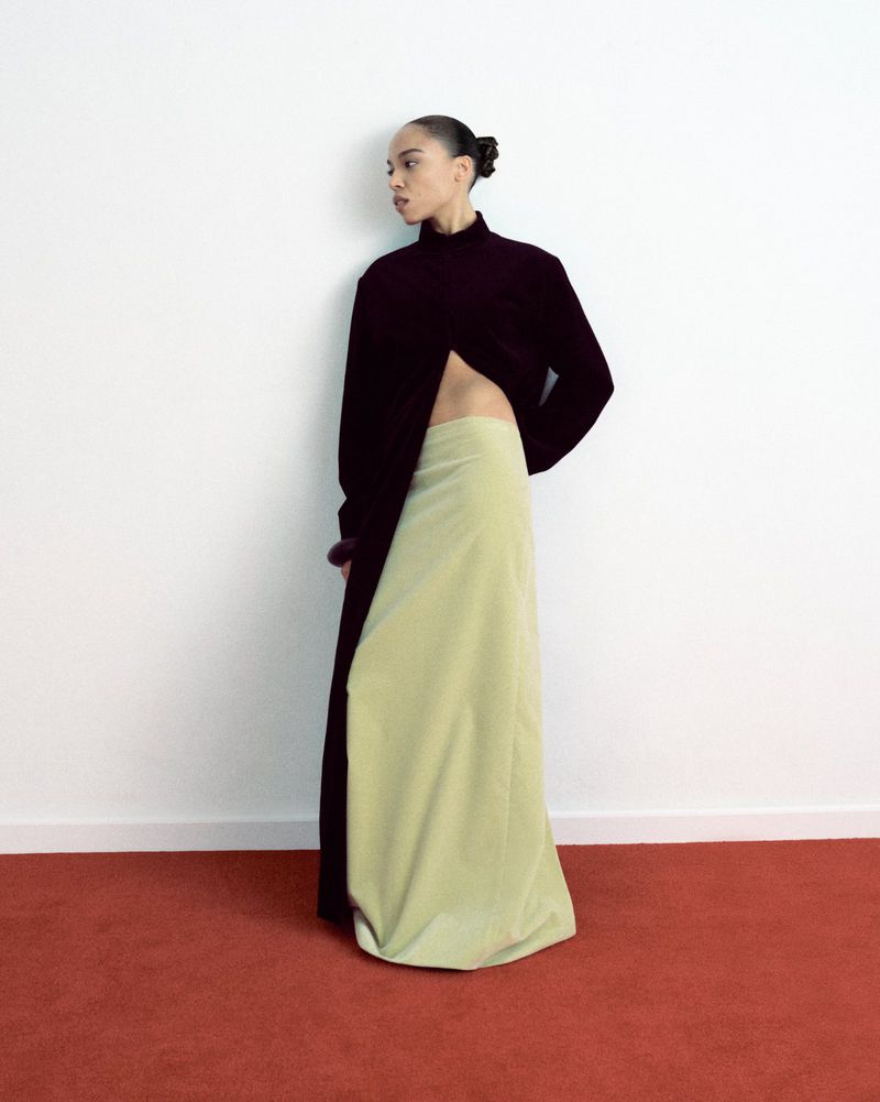 Fashionable woman in a split black top and light green maxi skirt poses against a white wall with a red carpet floor.