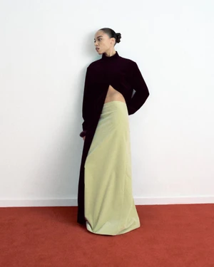 Model Zsela standing against a white wall wearing a dark split high-neck top and a pale green maxi skirt on a red carpet floor.