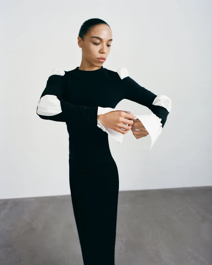 Model Zsela wearing a long black dress with white sculptural sleeve details against a plain white studio background.