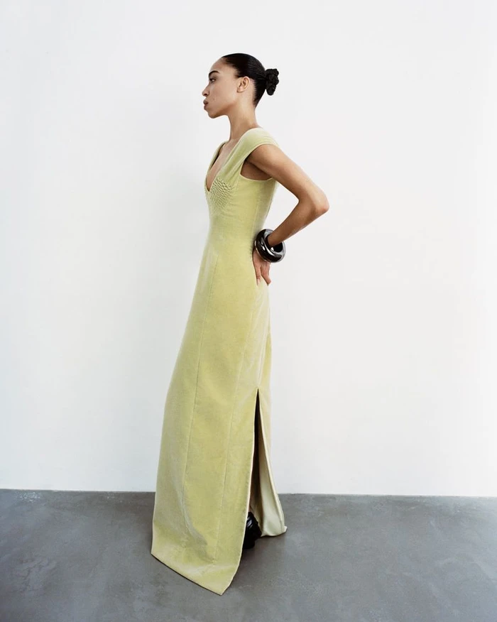 Model Zsela wearing a sleeveless pale yellow floor-length dress and chunky black bracelet against a white studio background.