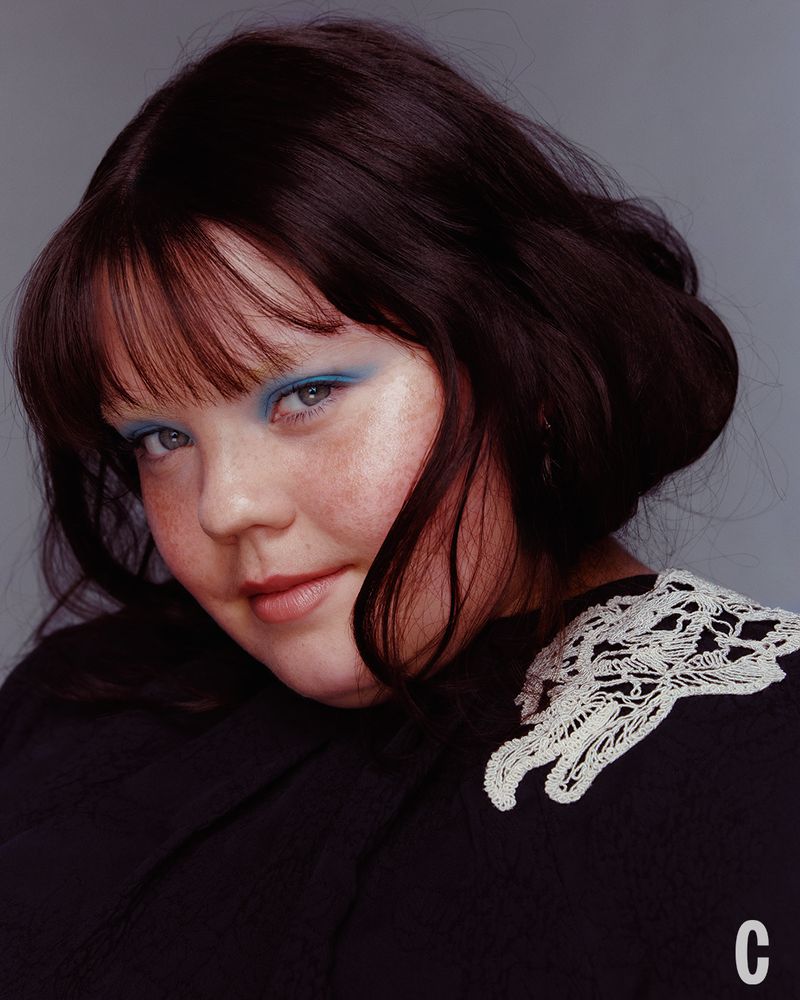 Portrait of a woman with dark bob hair, blue eye shadow, and black top with white lace collar against a grey background