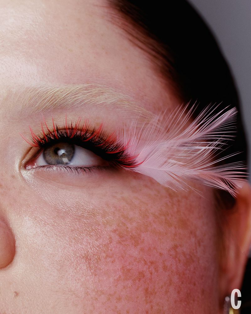 Close-up of a person's eye with pink and red eyelashes and feather extensions against a neutral background