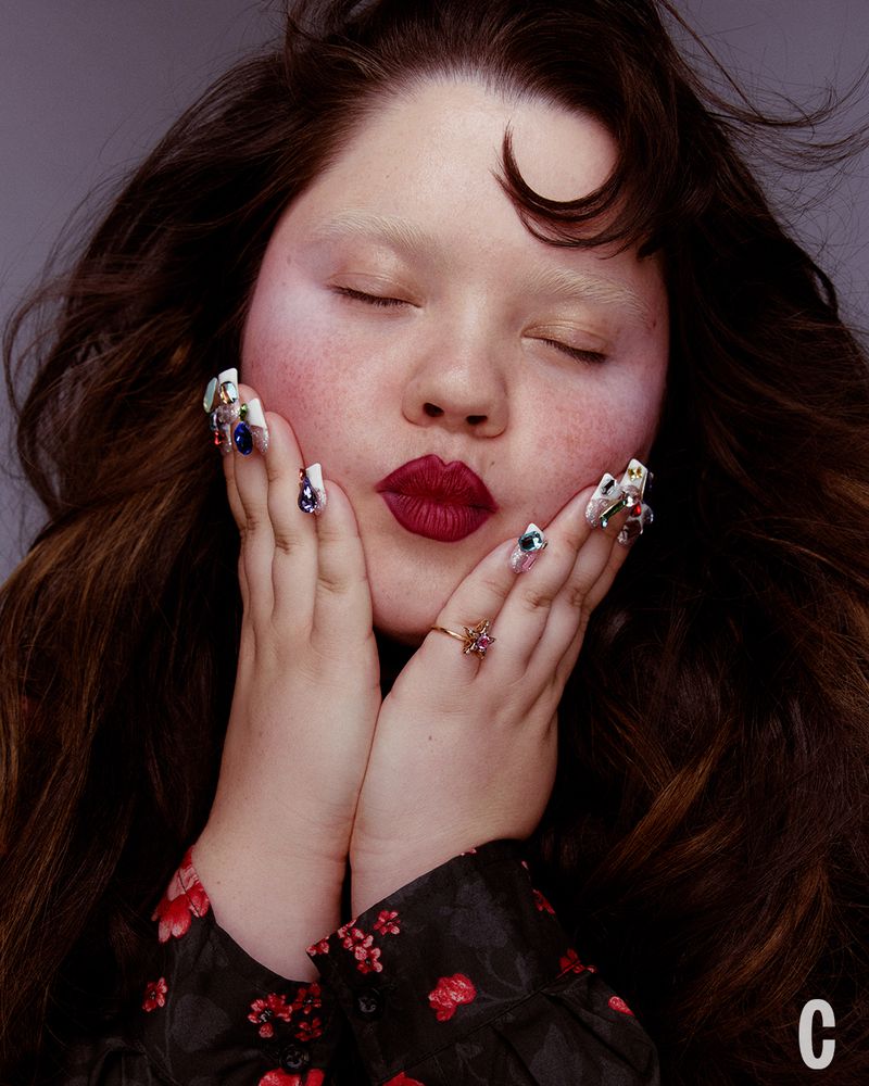 Close-up beauty portrait of a woman with dark hair, bold red lips, and gemstone-embellished long nails framing her face.