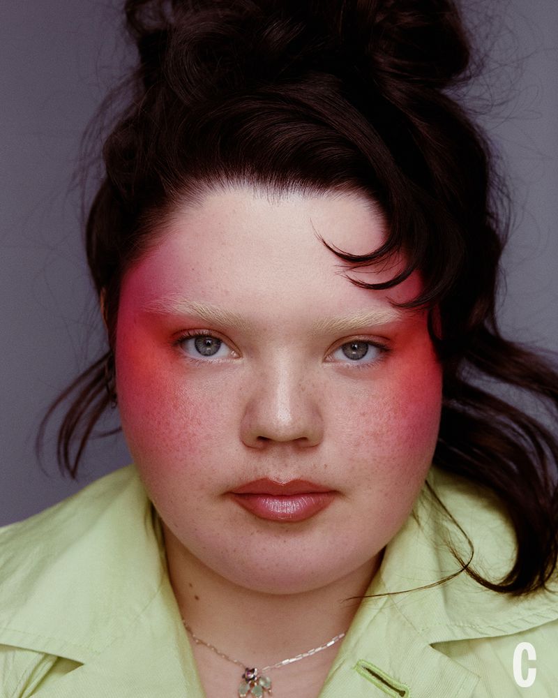 Close-up portrait of a woman with dramatic pink blush wearing a pale green jacket against a grey background